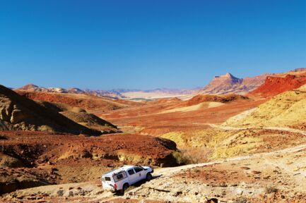Geländewagen fährt durch eine rote Wüstenlandschaft in Namibia mit Bergen im Hintergrund